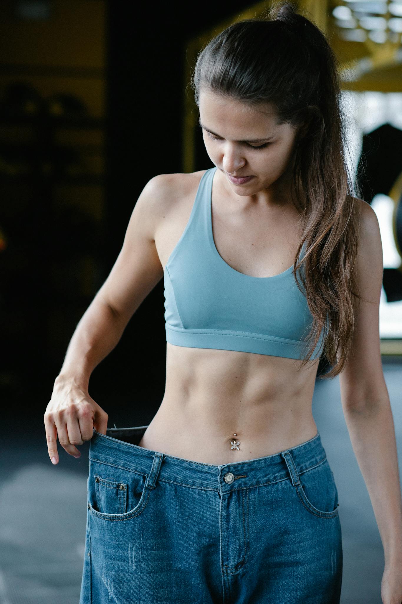 Fit woman showing weight loss success by wearing oversized jeans in gym.